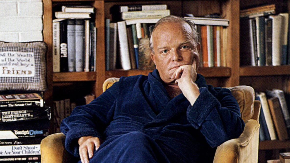 Truman Capote at home in blue robe seated in yellow armchair before a bookshelf